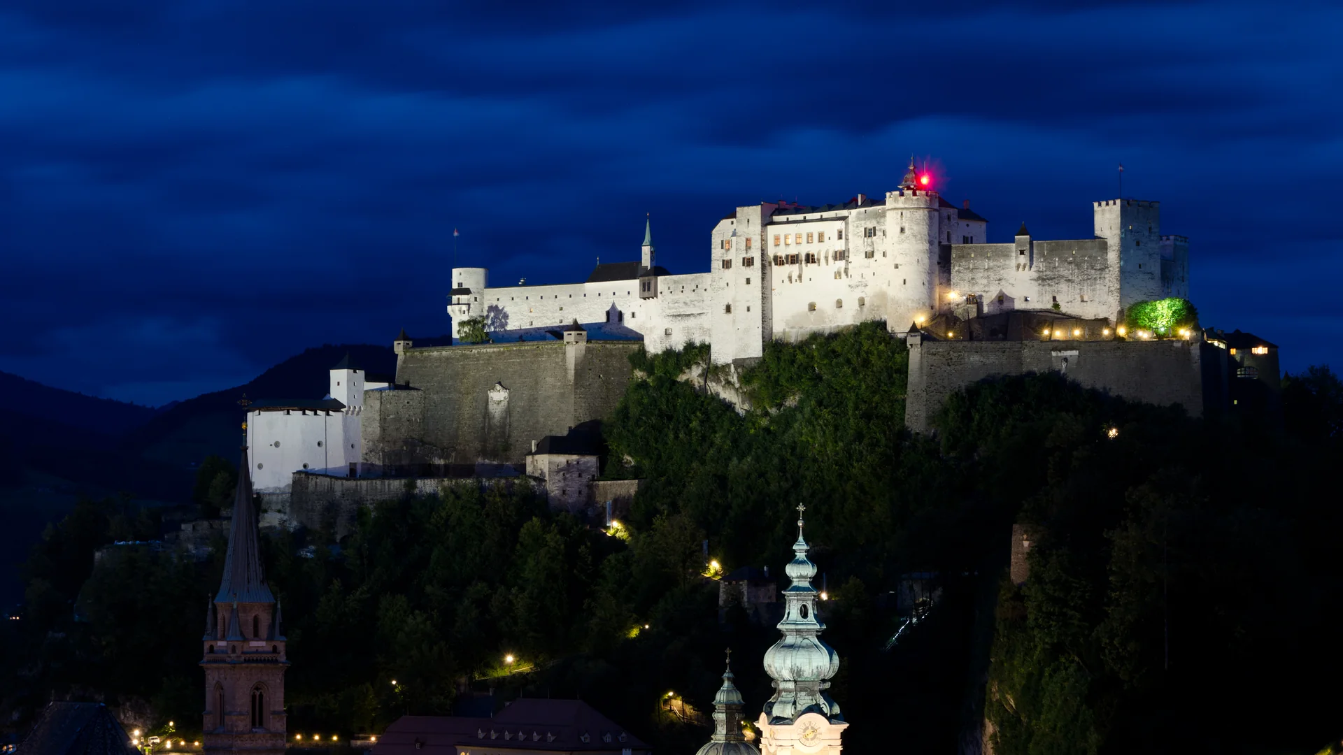 Hohensalzburg Fortress lit white on its hilltop against a deep blue dusk sky, with the green domes and spires of Salzburg's Old Town in the foreground