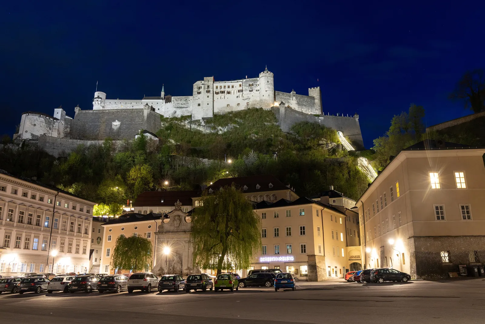 Hohensalzburg Fortress floodlit above a floodlit square in the Old Town at night, with parked cars and a tree in front