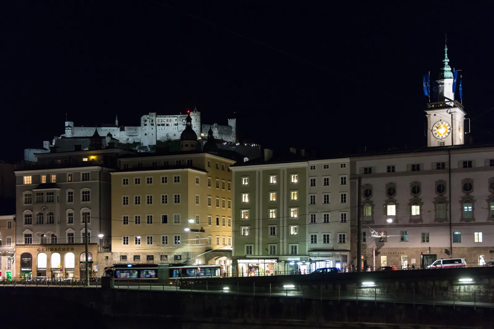 Houses along the Salzach embankment lit at night, with the Old Town clock tower on the right and the fortress glowing above the rooftops