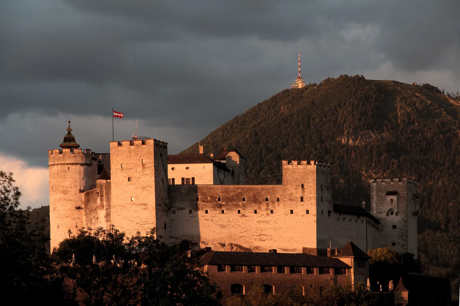 The towers and walls of Hohensalzburg Fortress lit orange by low sun, with the wooded Gaisberg hill rising behind